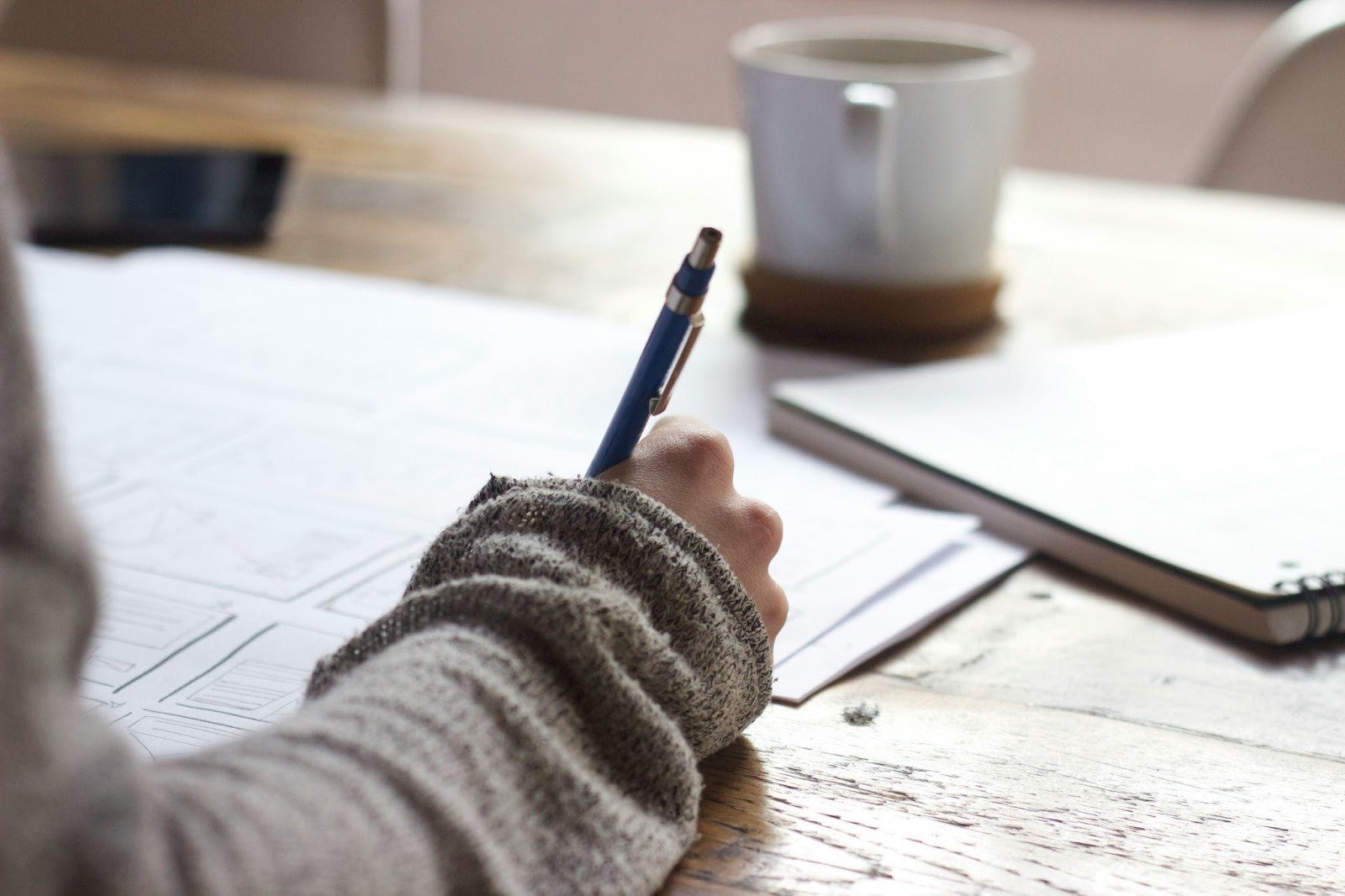 Hand holding a pen writing on paper at a wooden desk with a coffee mug and notebook in the background.
