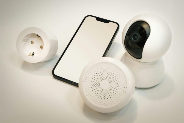 Smartphone surrounded by smart home devices including a security camera, smart plug, and round speaker sensor on a white surface.
