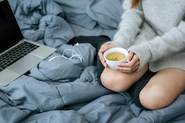 Person in a cozy sweater sitting on a bed holding a cup of herbal tea beside an open laptop and earphones.